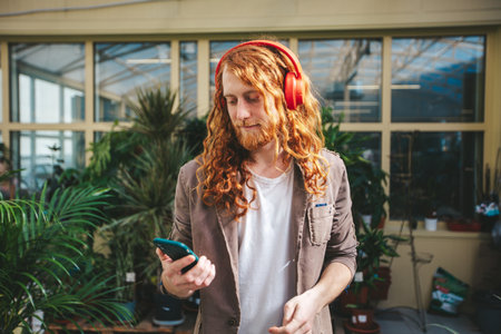 Young man with long red hair and beard wearing headphones and browsing his smartphone surrounded by plants in a luminous greenhouseの写真素材