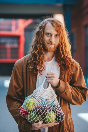 A man is holding a bag of fruit in his hand. The bag is made of a mesh material and contains a variety of fruits, including apples, avocado and oranges.の写真素材