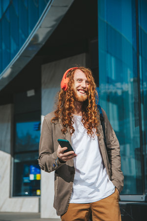 Happy business man with long curly red hair wearing headphones and listen to music near glass building, using technology and urban lifestyle concept.の写真素材