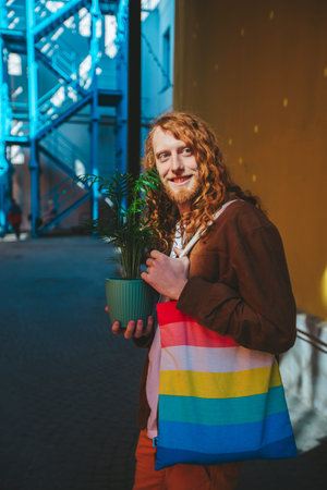 Happy young man with ginger hair and beard, holding a potted plant and a colorful bag, savoring a sunny day in the urban environmentの写真素材