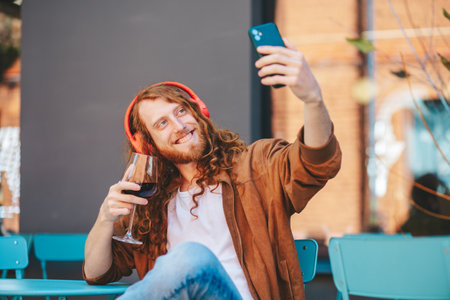 Young freelancer with headphones is taking a selfie with his smartphone while drinking a glass of wine, sitting at a table on a restaurants terraceの写真素材