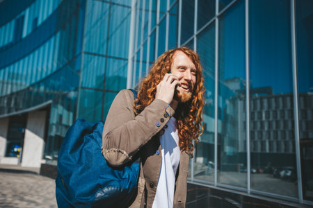 Young man with long red hair and a backpack is talking on his phone and smiling as he walks near a modern glass university buildingの写真素材