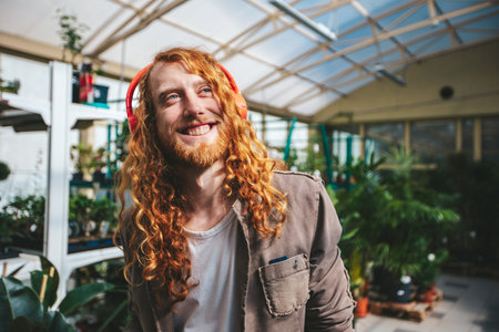 Young red hair man with beard is enjoying music over his headphones while working inside a greenhouse, surrounded by plants and flowersの写真素材