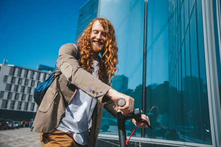 Young man with long red hair and a trendy beard happily rides an electric scooter through a bustling city center on a sunny day, showcasing eco friendly urban commutingの写真素材