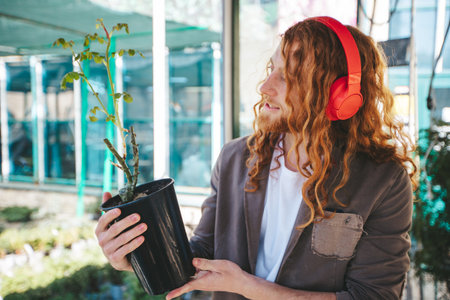 Nursery worker holding a rose plant in a pot while listening to music on his headphones, enjoying his job at the greenhouseの写真素材