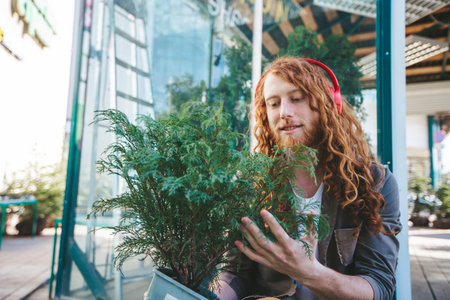 Red haired young adult man is holding a small tree while listening to music on his headphones. He is wearing casual clothing and smiling slightlyの写真素材