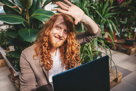 Young entrepreneur is shielding his eyes from the bright sun while working remotely on his laptop from a lush, plant filled greenhouseの写真素材