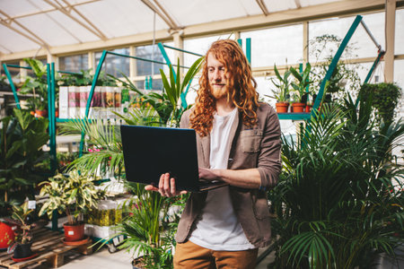 Small business owner manages his plant nursery with a laptop in a lush greenhouse, blending nature with technology for a productive and sustainable work environmentの写真素材