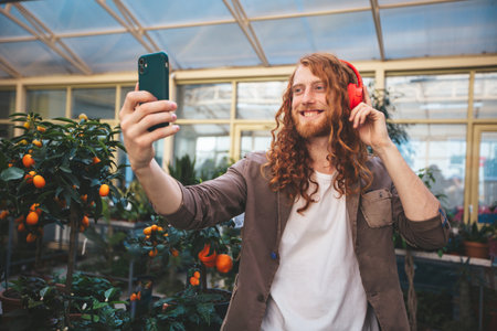 A man with red hair is taking a selfie in a greenhouse, wearing headphones and smiling to smartphone.の写真素材