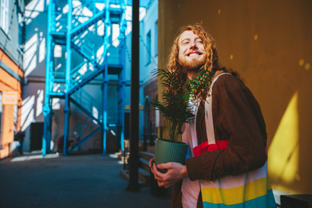 Young man with long red hair and a beard is holding a potted plant in an urban setting and smiling. He is wearing a brown jacket and carrying a colorful tote bagの写真素材