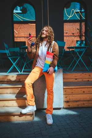 Young man with flowing hair stands confidently outside a coffee shop on a sunny day, clutching a reusable water bottleの写真素材