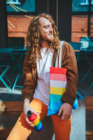 Fashionable man with flowing hair enjoying a drink outdoors in the city, using a colorful reusable water bottle for an eco friendly touchの写真素材