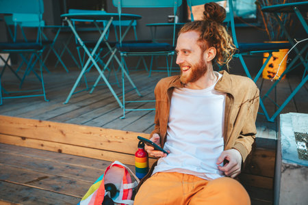 Young man in a colorful style happily laughs while texting on his phone at a trendy cafe terrace, embodying a carefree and positive urban lifestyleの写真素材