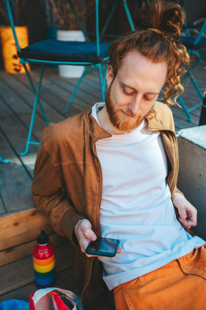 Young man with ginger hair and a beard, dressed in a brown jacket and orange pants, sits on a bar terrace while using his smartphoneの写真素材