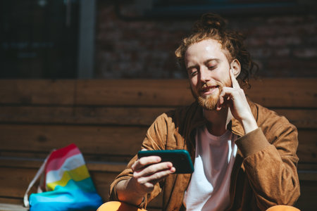 Happy young man with red hair and a beard enjoys using his smartphone outdoors on a sunny day, with an lgbt pride flag representing freedom and support in the backgroundの写真素材