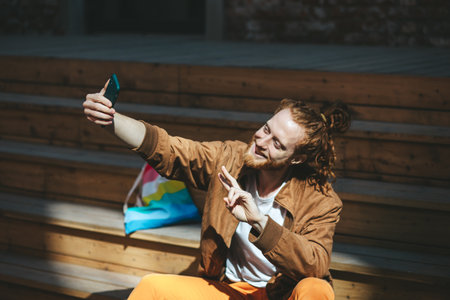 Young man with a beard and long red hair tied in a bun is taking a selfie with his smartphone while sitting on wooden steps and making the peace sign with his handの写真素材