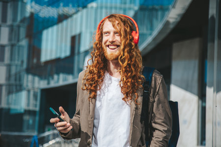 Happy business man with long curly red hair wearing headphones and listen to music near glass building, using technology and urban lifestyle concept.の写真素材