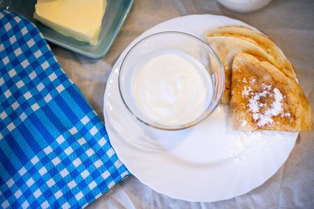 Pancakes on a white plate with powdered sugar and sour cream, serving for Breakfast, blue tablecloth