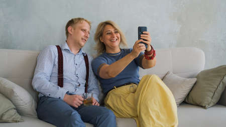 Smiling Mature Mother with Adult Son Taking Selfies on the Smartphone while Sitting on Sofa at Home. Bonding and Family Reunion, Mothers Day Conceptの写真素材