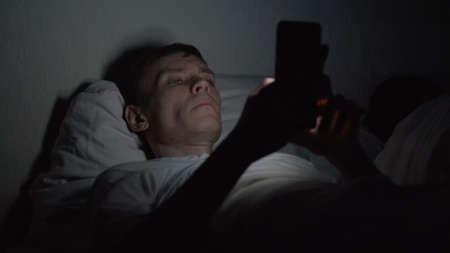 Young Man Lying in Bed and Using Smartphone at Late Night, News and Social Media Addiction. Insomnia and Sleep Disorder Conceptの写真素材