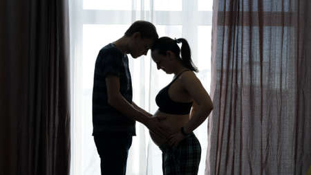 Silhouettes of Pregnant Couple Standing by Window, Male Touching Pregnant Belly of his Wife. Young Family Preparing for Newbornの写真素材