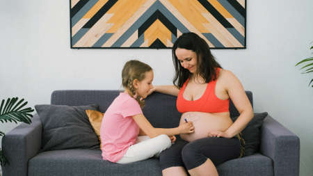 Little Girl Draws the Heart on Belly of Pregnant Mother with Felt-tip Pen while Sittin on Sofa at Homeの写真素材