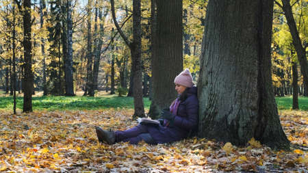Young trendy woman reading a book and drinking coffee while leaning on tree in scenic autumn parkの写真素材