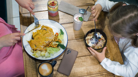 Little girl with mother having healthy breakfast at cafe or restaurant. Young woman eating vegan chickpea omelet and her daughter eating blueberry smoothie bowl. View from the topの写真素材