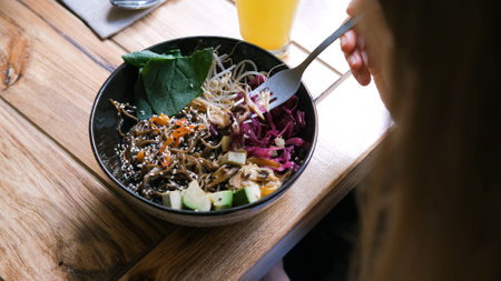 Young woman having poke bowl with buckwheat noodles and vegetables and orange shake on a lunch, slow...