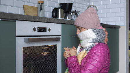 Young woman warming her hands while sitting on the floor by oven in the kitchen at homeの写真素材