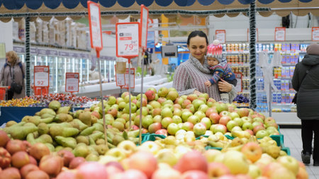 Young woman choosing fresh apples while holding baby in hands at store. NOVOSIBIRSK, RUSSIA, NOVEMBER 21, 2022のeditorial素材