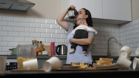 Tired woman lulling the child and trying to drink coffee in messy kitchen. Single mother conceptの写真素材