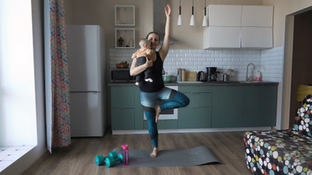 Young mother holding baby girl in hands and practicing yoga in the kitchenの写真素材