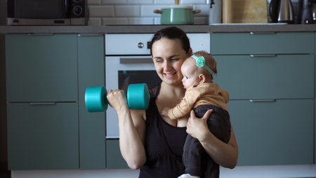 Happy young mother holding baby in hands while training with dumbbells in the kitchen at homeの写真素材
