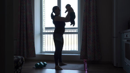 Silhouettes of sporty young mother holding baby girl in hands and doing squatsの写真素材