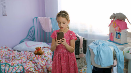 Little girl in pink dress grimacing while taking selfies on her smart phone in messy roomの写真素材