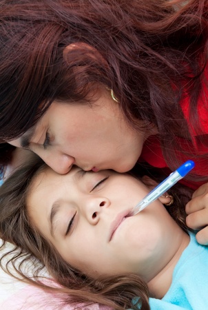 Latin mother kissing her sick daughter who has a thermometer in her mouthの写真素材