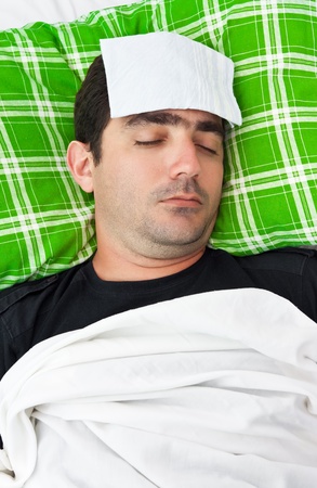 Sick hispanic man laying in bed with a tissue in his forehead to relieve him from fever or headacheの写真素材