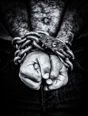 Dramatic black and white image of the hands of a shirtless  man chained with an iron chain and a padlock  emerging from a black background の写真素材
