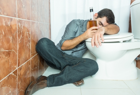 Drunk hispanic man sleeping over the toilet bowl and holding a whisky bottleの写真素材