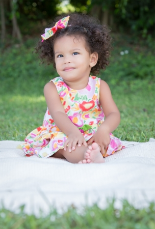 Funny small latin girl with a colorful dress sitting on a green grass parkの写真素材
