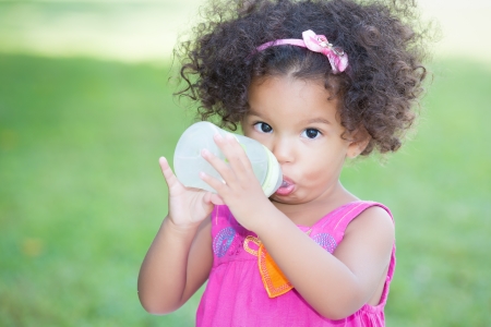 Cute latin girl drinking from a baby bottle with a green grass backgroundの写真素材