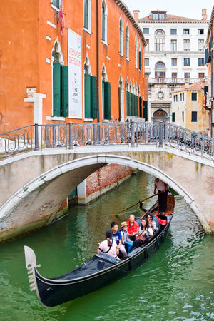 Gondola carrying tourists under a bridge on a narrow canal sidelined by old palacesのeditorial素材