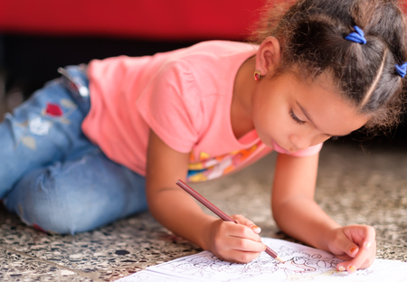 Cute multiracial small girl drawing on a coloring book while lying on the floorの写真素材