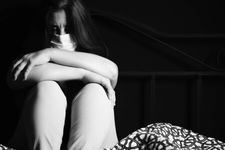 Sad and stressed woman wearing a face mask to avoid disease contagion - Black and white portrait sitting on a bedの写真素材