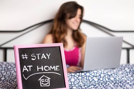 Teenager in her bedroom using a laptop computer during a quarantine with a Stay At Home sign - Selective focus, focused on the signの写真素材