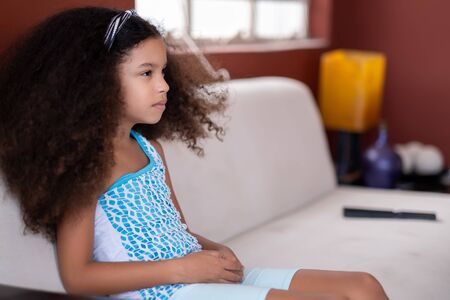 Cute multiracial small girl with beautiful curly hair watching TV at homeの写真素材
