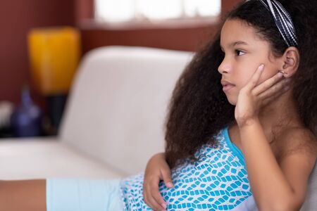 Cute multiracial small girl with beautiful curly hair watching TV at homeの写真素材