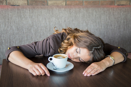 Beautiful tired young woman falling asleep while drinking coffee	at a cafeの写真素材