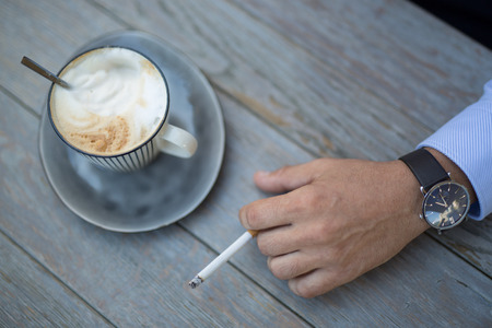 Closeup shot of a cup of coffee and a mans hand with a cigarette daytimeの写真素材